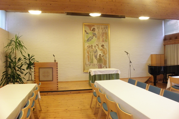 Pöytiä ja tuoleja seurakuntasalissa, piano ja seinällä tekstiiliteos.